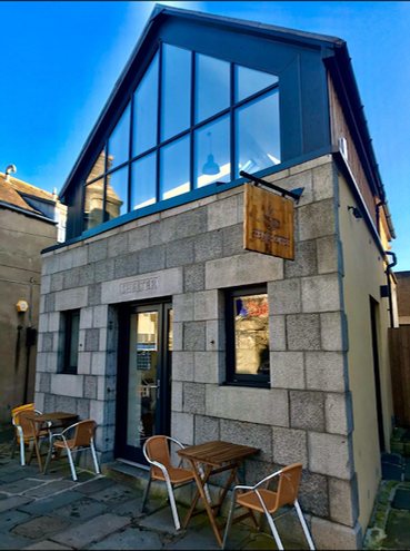 The front of Shelter Coffee: a granite building with a tall glass gable, a wooden hanging sign and tables and chairs outside
