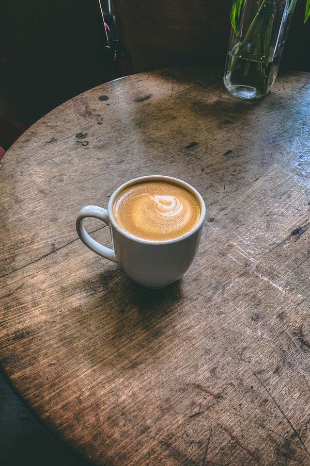 A small white cup of coffee with a creamy top on a worn round wooden table